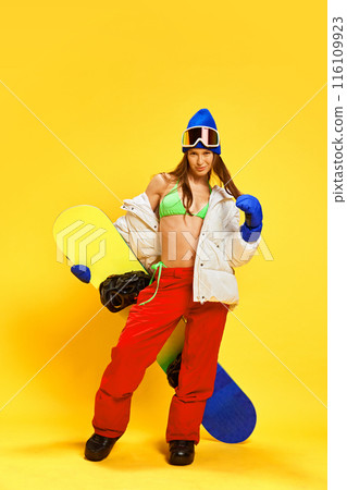 Athletic woman in green bikini top and winter gear, showcasing her toned body and adventurous spirit against bright yellow studio background 116109923