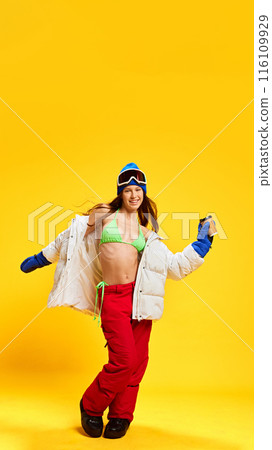 Happy woman in unique combination of winter and summer clothing, enjoying vacation vibes against bright yellow studio background 116109929