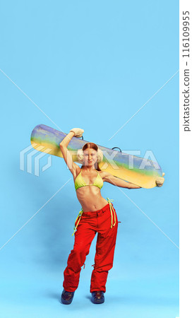 Body confidence in winter. Beautiful tanned young girl in bikini and winter pants posing with snowboarder against blue studio background Body confidence in winter. Beautiful tanned young girl in bikini and winter pants posing with snowboarder against blue studio background 116109955