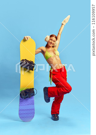 Full-length portrait of happy young smiling girl wearing red pants and green bikini top, poses with snowboard against blue studio background 116109957