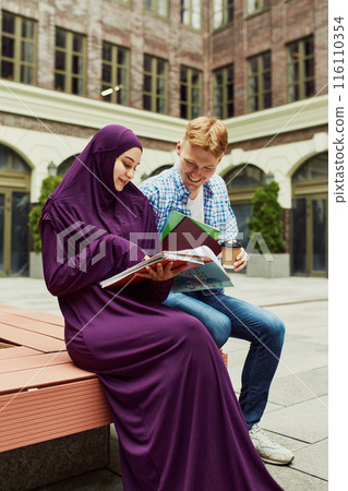 Smiling friends, students, Muslim girl and Caucasian boy sitting outside the campus and preparing for lesson together. Smiling friends, students, Muslim girl and Caucasian boy sitting outside the campus and preparing for lesson together. 116110354