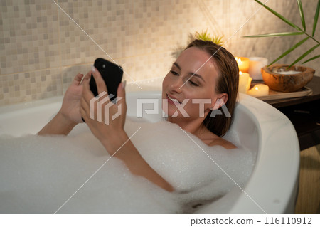 Woman relaxing in a bubble bath at night. She is using her smartphone to browse the internet or watch a video. The bathroom is lit by candles and there is a plant in the background. 116110912