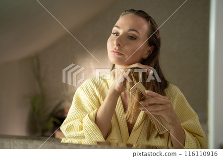 Woman combing her hair in front of a mirror. She is wearing a yellow robe and appears to be in the bathroom. 116110916
