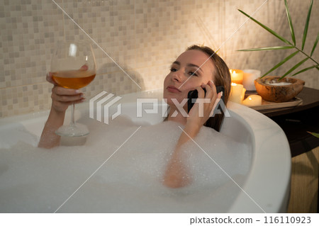 A woman is seen enjoying a relaxing evening in the bathtub filled with bubbles. She is holding a glass of wine and talking on the phone. The warm glow of candlelight creates a cozy atmosphere. 116110923