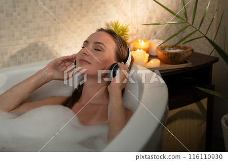 A woman is enjoying a relaxing evening in a bubble bath. She is listening to music with headphones on. The bathroom is dimly lit with candles, and the woman looks serene and content. 116110930
