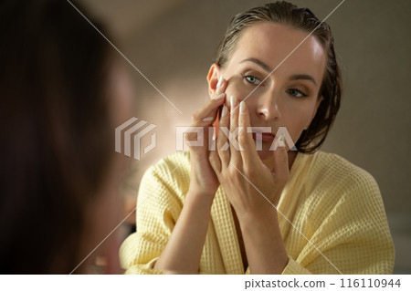 Woman looking in a mirror, focusing on the pimples on her face. She looks concerned and worried about her skin. 116110944