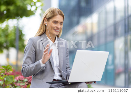 Professional woman in a grey suit participating in a video conference call on her laptop while sitting outdoors. She is wearing earphones and smiling, communicating in a business setting. Professional woman in a grey suit participating in a video conference call on her laptop while sitting outdoors. She is wearing earphones and smiling, communicating in a business setting. 116111067