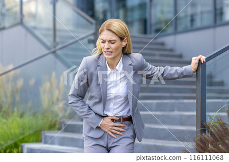 A businesswoman in a grey suit experiencing severe stomach pain while standing outside an office building. She is holding her abdomen and showing discomfort. The image highlights feelings of pain A businesswoman in a grey suit experiencing severe stomach pain while standing outside an office building. She is holding her abdomen and showing discomfort. The image highlights feelings of pain 116111068