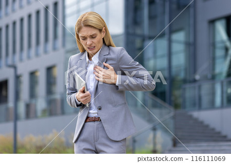 A stressed businesswoman clutches her chest in pain, standing outside a modern office building. She is dressed in a gray suit and holding a tablet, indicating a professional setting health concern. A stressed businesswoman clutches her chest in pain, standing outside a modern office building. She is dressed in a gray suit and holding a tablet, indicating a professional setting health concern. 116111069