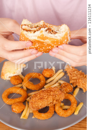 Caucasian man eats half-eaten hamburger, potato chips, onion rings and chicken pieces in gray plate on wooden table 116111234