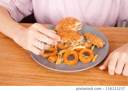 Caucasian man eating fried chicken wings, potato chips, onion rings and chicken wings by hand from a gray plate on a wooden table 116111237