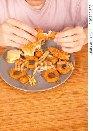 Caucasian man eating fried chicken wings, potato chips, onion rings and chicken wings by hand from a gray plate on a wooden table Caucasian man eating fried chicken wings, potato chips, onion rings and chicken wings by hand from a gray plate on a wooden table 116111263