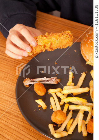 Caucasian man eating fried chicken wings, potato chips, onion rings and chicken wings by hand from a gray plate on a wooden table Caucasian man eating fried chicken wings, potato chips, onion rings and chicken wings by hand from a gray plate on a wooden table 116111339