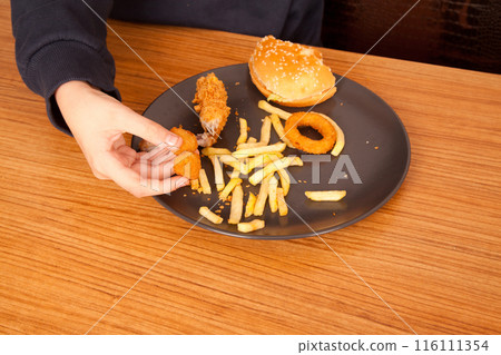 Caucasian man eating fried chicken wings, potato chips, onion rings and chicken wings by hand from a gray plate on a wooden table 116111354