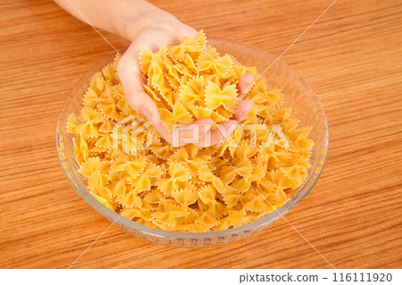 Uncooked raw healthy and pasta macaroni hand held by a Caucasian man, on wooden table in glass plate 116111920