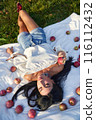 Relaxed pretty woman lying with apples on white rug on grass in garden. View from above of calm Caucasian woman in casual clothes resting on ground, surrounded by apples at sunny day. Picnic concept. 116112432
