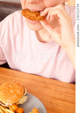 Caucasian man eating fried chicken wings, potato chips, onion rings and chicken wings by hand from a gray plate on a wooden table 116112734