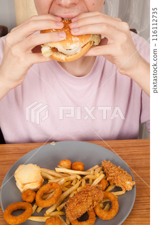 Caucasian man eats half-eaten hamburger, potato chips, onion rings and chicken pieces in gray plate on wooden table 116112735