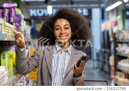 Smiling woman in a suit shopping at a supermarket aisle. She is choosing products while holding a smartphone. The image captures everyday life, consumerism, and grocery shopping experiences. Smiling woman in a suit shopping at a supermarket aisle. She is choosing products while holding a smartphone. The image captures everyday life, consumerism, and grocery shopping experiences. 116112780