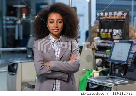 A confident supermarket cashier manager with curly hair in business attire standing with arms crossed at a checkout counter. 116112782