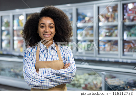 A cheerful supermarket employee wearing an apron and plaid shirt stands confidently with arms crossed in front of the refrigerated section. She exudes friendliness and professionalism 116112788