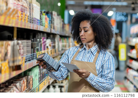 Store employee in apron checking inventory and scanning a product with a digital tablet in the beauty supply aisle. Organized shelves with various cosmetic products. Focus on detailed work in retail Store employee in apron checking inventory and scanning a product with a digital tablet in the beauty supply aisle. Organized shelves with various cosmetic products. Focus on detailed work in retail 116112798