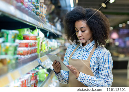 A grocery store employee in an apron checks the inventory on the shelves using a tablet. The worker appears focused and attentive, ensuring the store is well-stocked and organized for customers. A grocery store employee in an apron checks the inventory on the shelves using a tablet. The worker appears focused and attentive, ensuring the store is well-stocked and organized for customers. 116112801