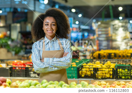 A cheerful grocery store employee with a big smile stands in front of the colorful organic produce section. The worker is wearing an apron and a checkered shirt A cheerful grocery store employee with a big smile stands in front of the colorful organic produce section. The worker is wearing an apron and a checkered shirt 116112803