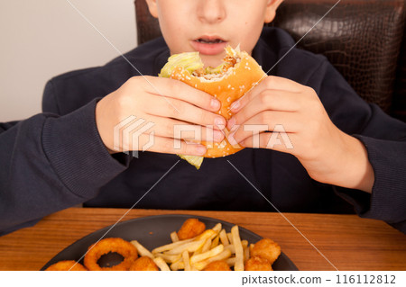 Caucasian man eats half-eaten hamburger, potato chips, onion rings and chicken pieces in gray plate on wooden table Caucasian man eats half-eaten hamburger, potato chips, onion rings and chicken pieces in gray plate on wooden table 116112812
