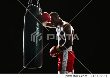 Feeling tired after intensive training. Muscular African man with strong body, boxer leaning on punching bag isolated on black background 116113331