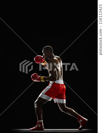 Concentrated African-American man, boxer with sweating shirtless body training, punching isolated on black background. Side view 116113423