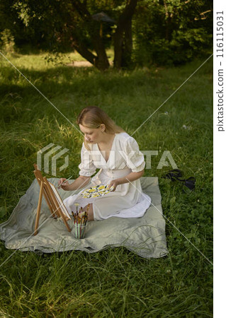 a beautiful young artist in a white dress paints a picture on the river bank a beautiful young artist in a white dress paints a picture on the river bank 116115031