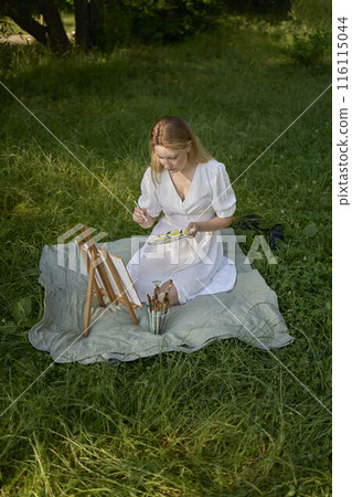 a beautiful young artist in a white dress paints a picture on the river bank a beautiful young artist in a white dress paints a picture on the river bank 116115044