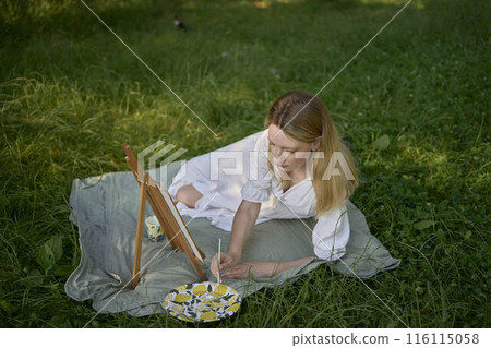a beautiful young artist in a white dress paints a picture on the river bank 116115058