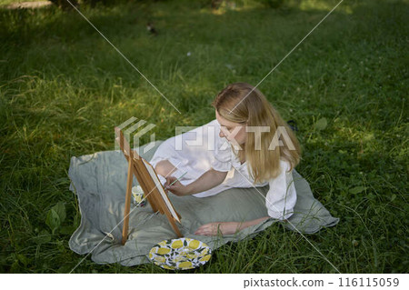 a beautiful young artist in a white dress paints a picture on the river bank a beautiful young artist in a white dress paints a picture on the river bank 116115059