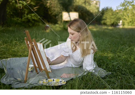 a beautiful young artist in a white dress paints a picture on the river bank a beautiful young artist in a white dress paints a picture on the river bank 116115060