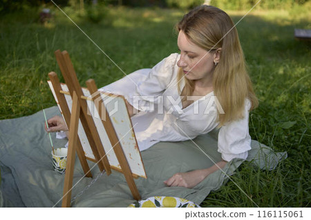 a beautiful young artist in a white dress paints a picture on the river bank a beautiful young artist in a white dress paints a picture on the river bank 116115061
