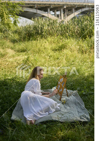 a beautiful young artist in a white dress paints a picture on the river bank 116115063
