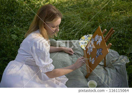 a beautiful young artist in a white dress paints a picture on the river bank a beautiful young artist in a white dress paints a picture on the river bank 116115064