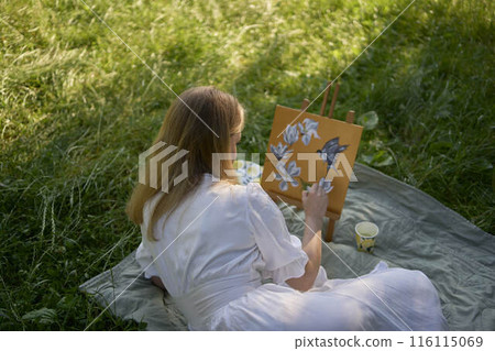 a beautiful young artist in a white dress paints a picture on the river bank 116115069