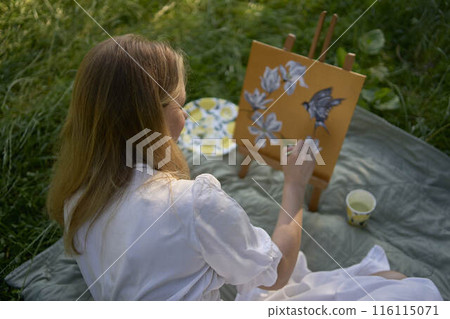 a beautiful young artist in a white dress paints a picture on the river bank a beautiful young artist in a white dress paints a picture on the river bank 116115071