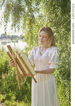 beautiful young artist in a white dress among the trees on the river bank 116115122
