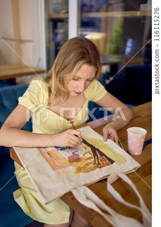 a beautiful young artist in a yellow dress paints a customized shopping bag a beautiful young artist in a yellow dress paints a customized shopping bag 116115276