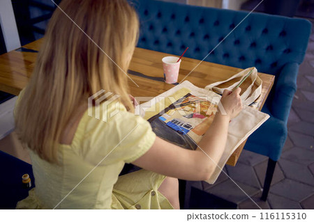 a beautiful young artist in a yellow dress paints a customized shopping bag a beautiful young artist in a yellow dress paints a customized shopping bag 116115310