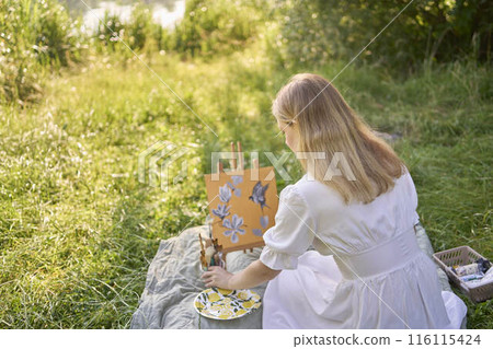 a beautiful young artist in a white dress paints a picture on the river bank 116115424