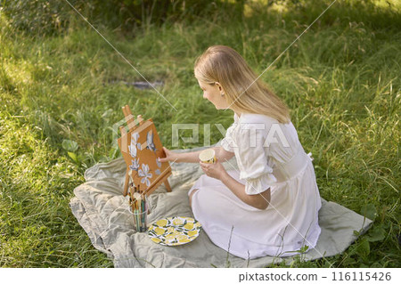 a beautiful young artist in a white dress paints a picture on the river bank 116115426