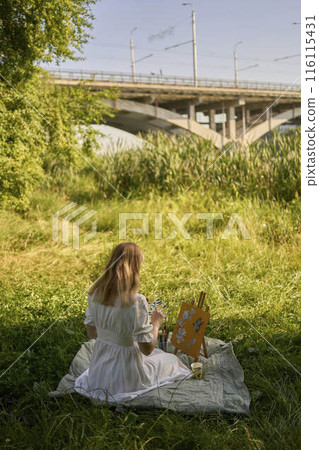 a beautiful young artist in a white dress paints a picture on the river bank a beautiful young artist in a white dress paints a picture on the river bank 116115431