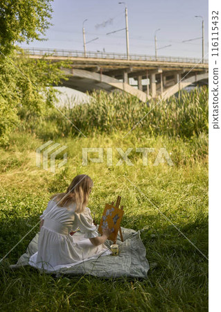 a beautiful young artist in a white dress paints a picture on the river bank a beautiful young artist in a white dress paints a picture on the river bank 116115432