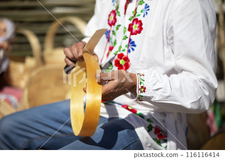 A Slavic man in an embroidered shirt makes products from birch bark. A Slavic man in an embroidered shirt makes products from birch bark. 116116414