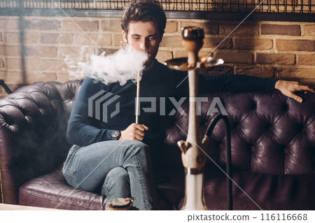 awesome brutal guy is sitting with crossed legs on the sofa and using the hookah awesome brutal guy is sitting with crossed legs on the sofa and using the hookah 116116668